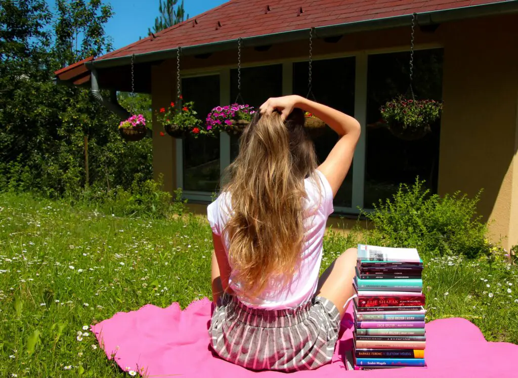A woman setting on the grass with a pile of books next to her.